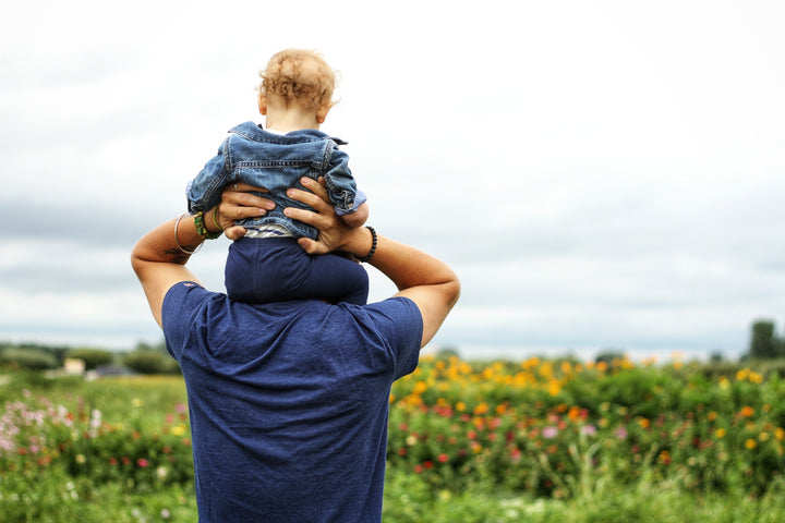 A man with his child on his shoulders, back facing the camera, walking in a field of wild flowers.